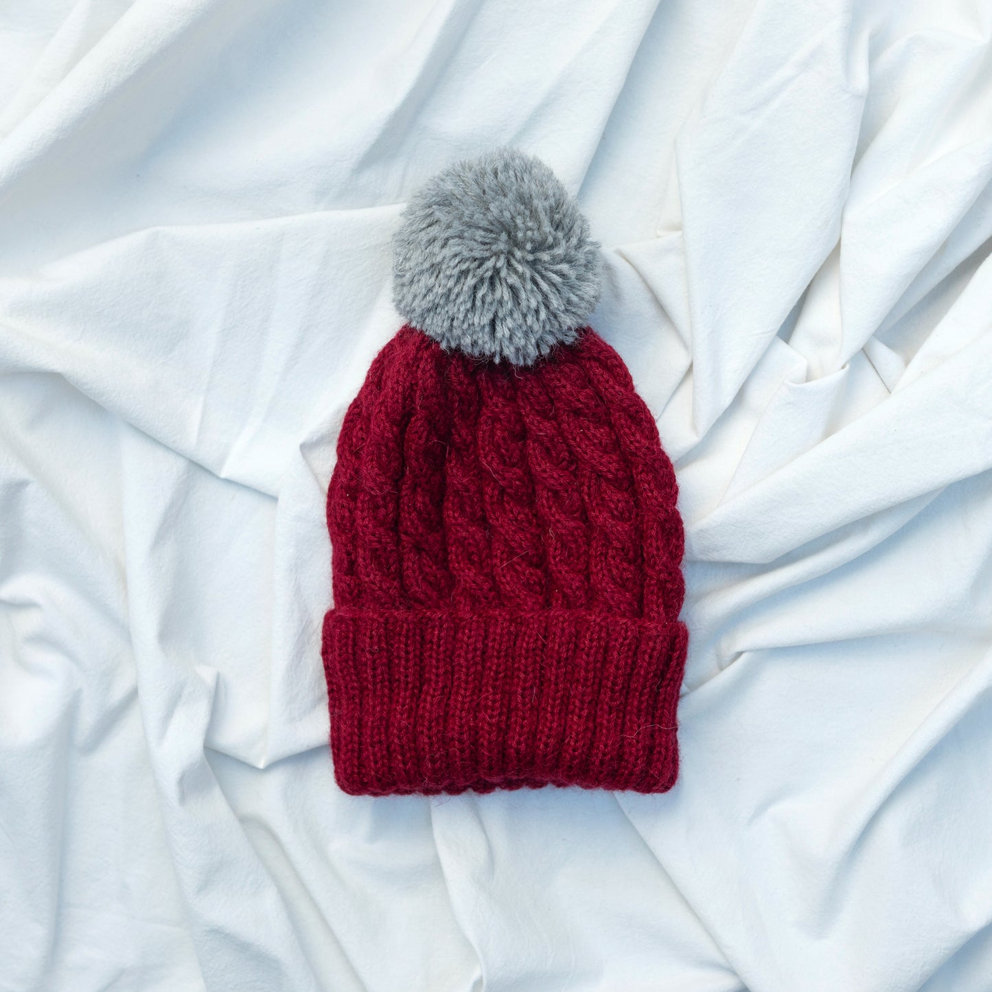 Burgundy coloured hand knitted cable beanie hat with grey pompom