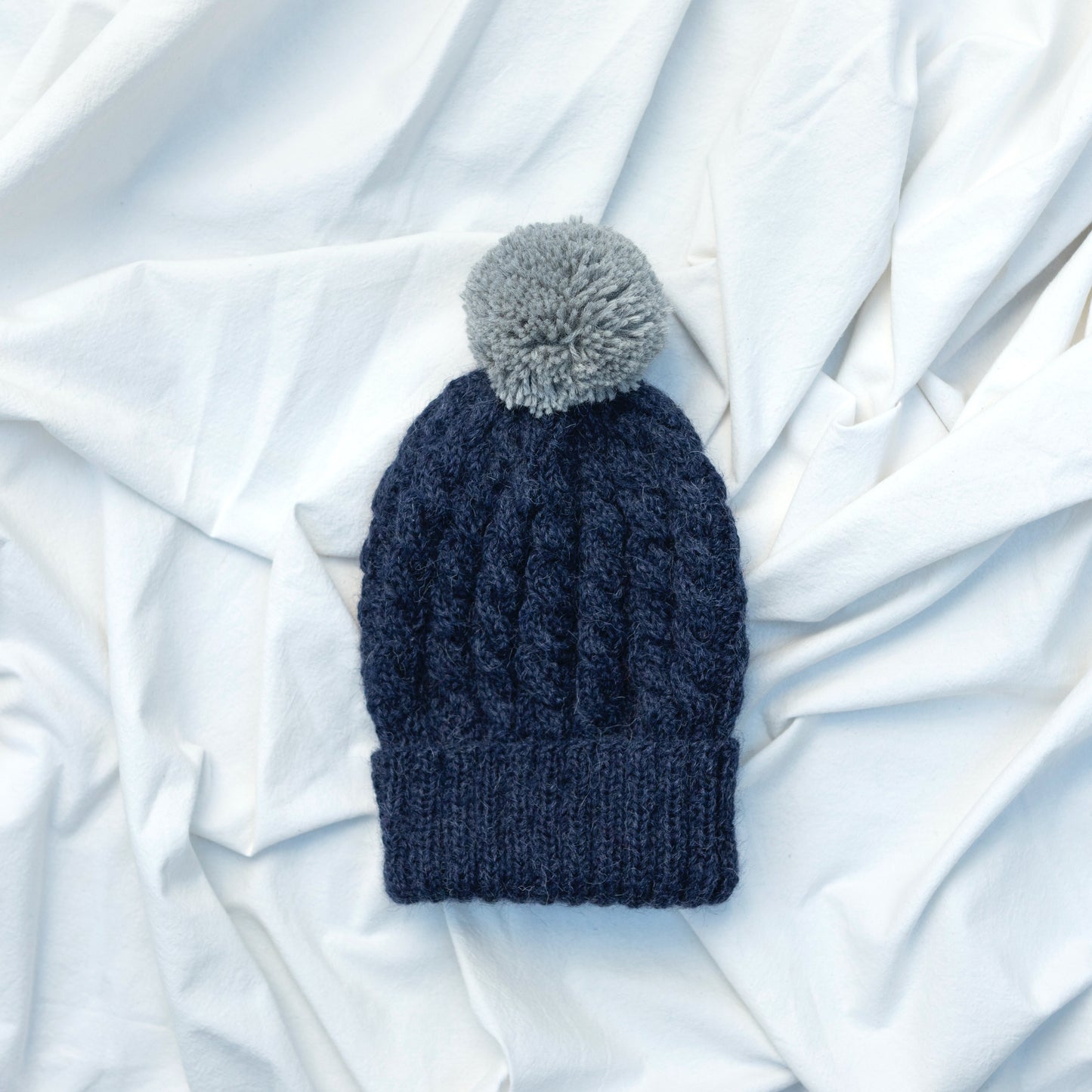 Dark blue coloured hand knitted cable beanie hat with grey pompom