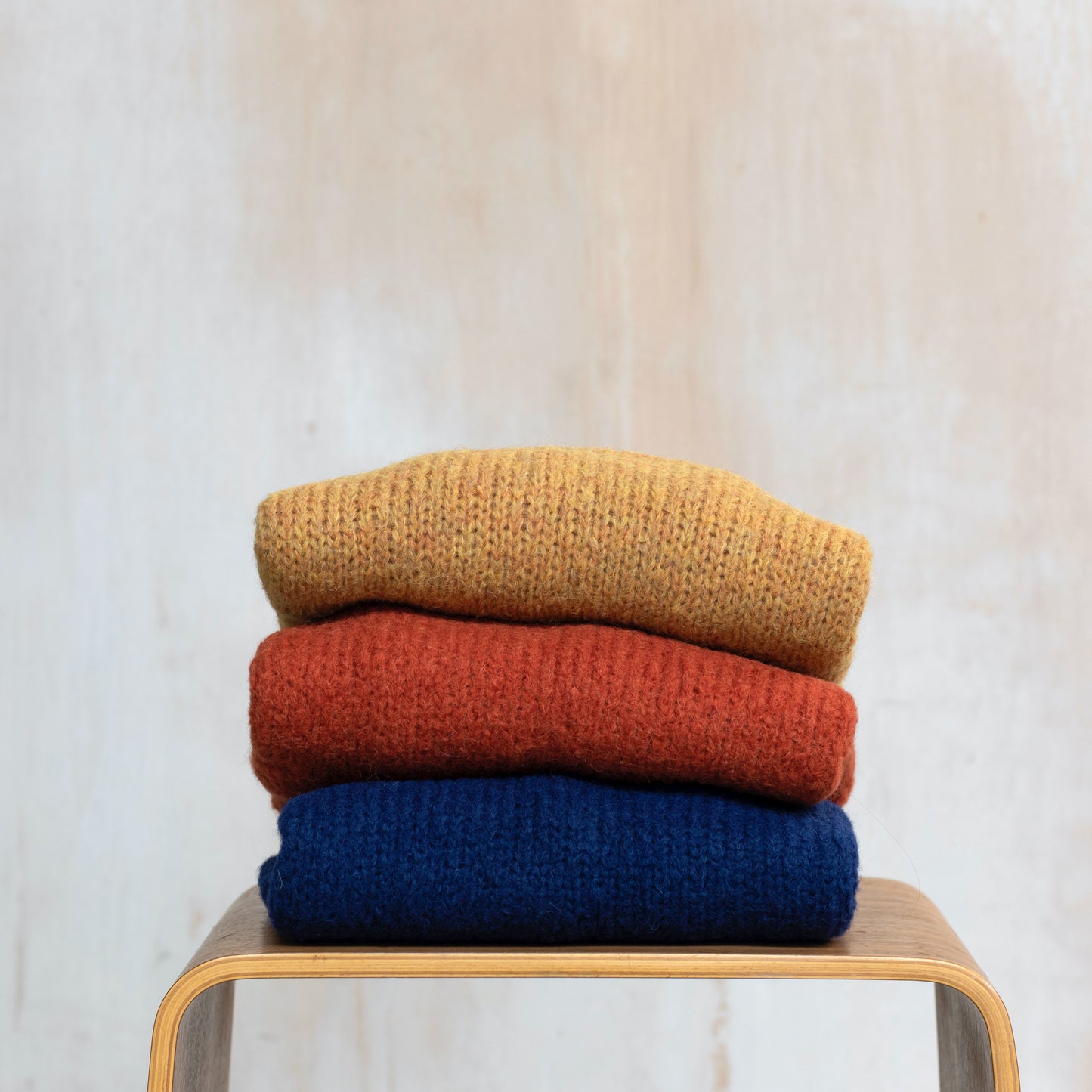 Three alpaca sweaters folded on a chair, yellow, orange and navy blue