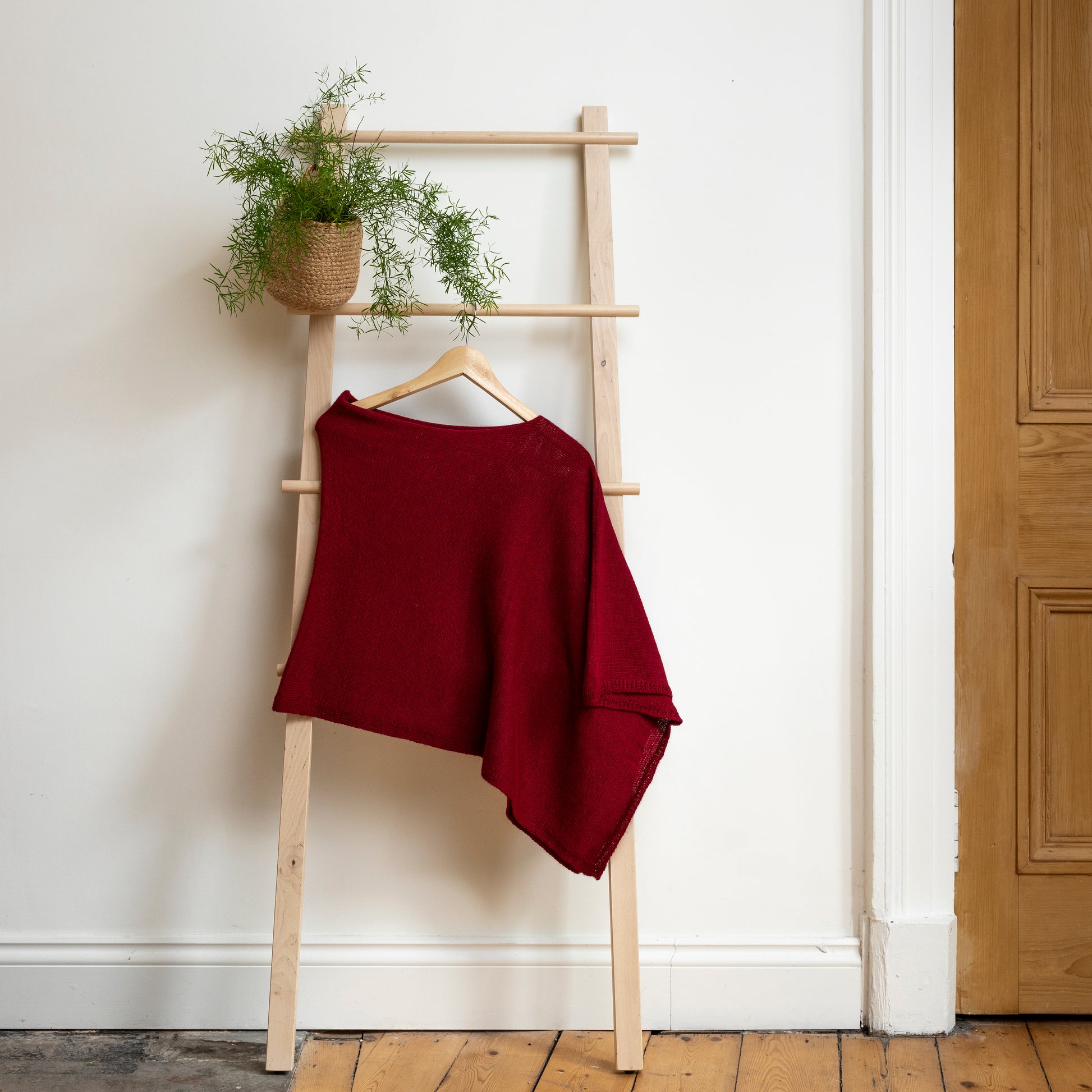 Asymmetrical burgundy luxury knitted poncho hanging on wooden ladder.