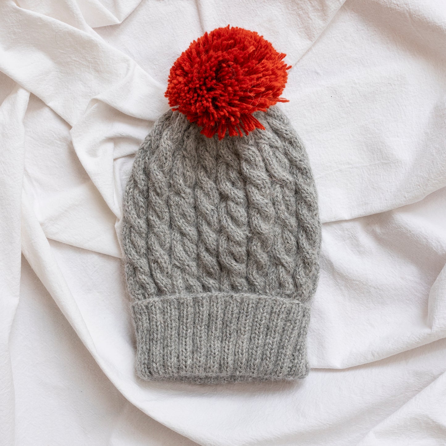 Woman wearing stone coloured hand-knitted cable beanie hat with red pom pom